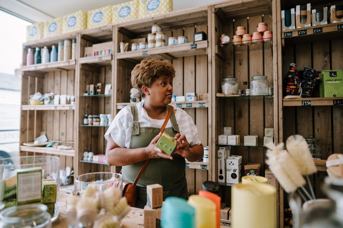 A shop owner in an apron restocking a shelf of refillable goods, mid-conversation with a customer.