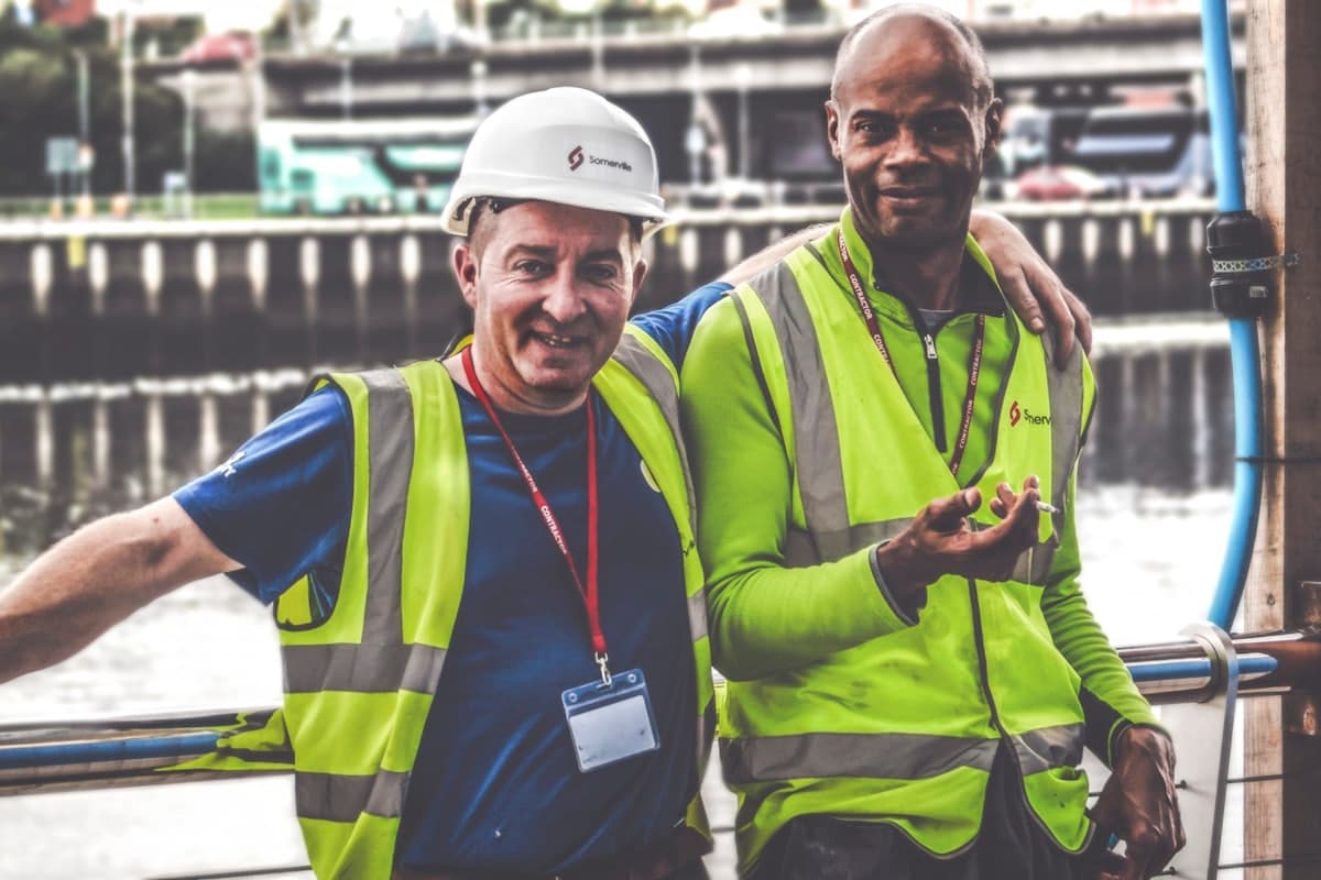 Two contractors in hi-vis on a job site, one with an arm around the other, both laughing.